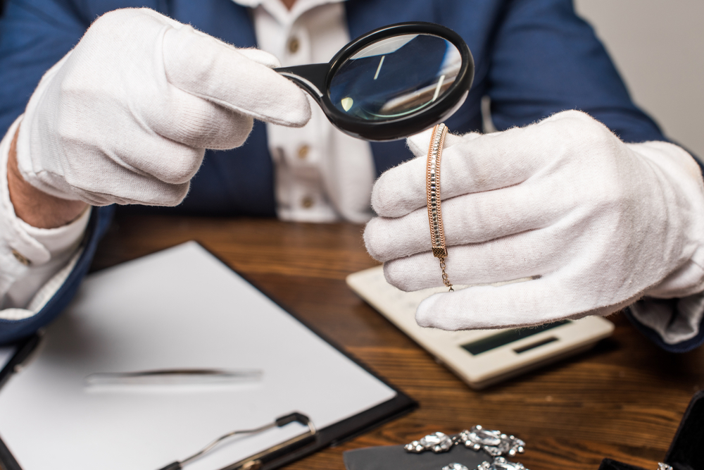 a jewelry appraiser wearing white gloves inspects a gold and diamond bracelet using a magnifying a glass